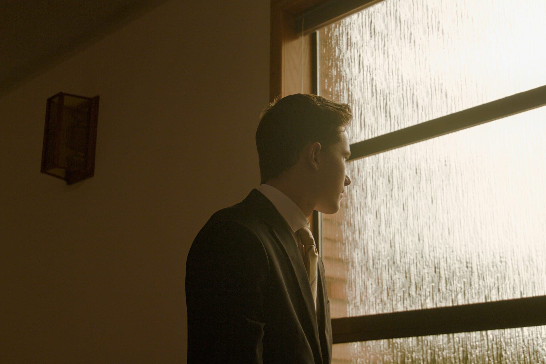 Man in a suit and tie in warm window light, looking out through rain-streaked glass