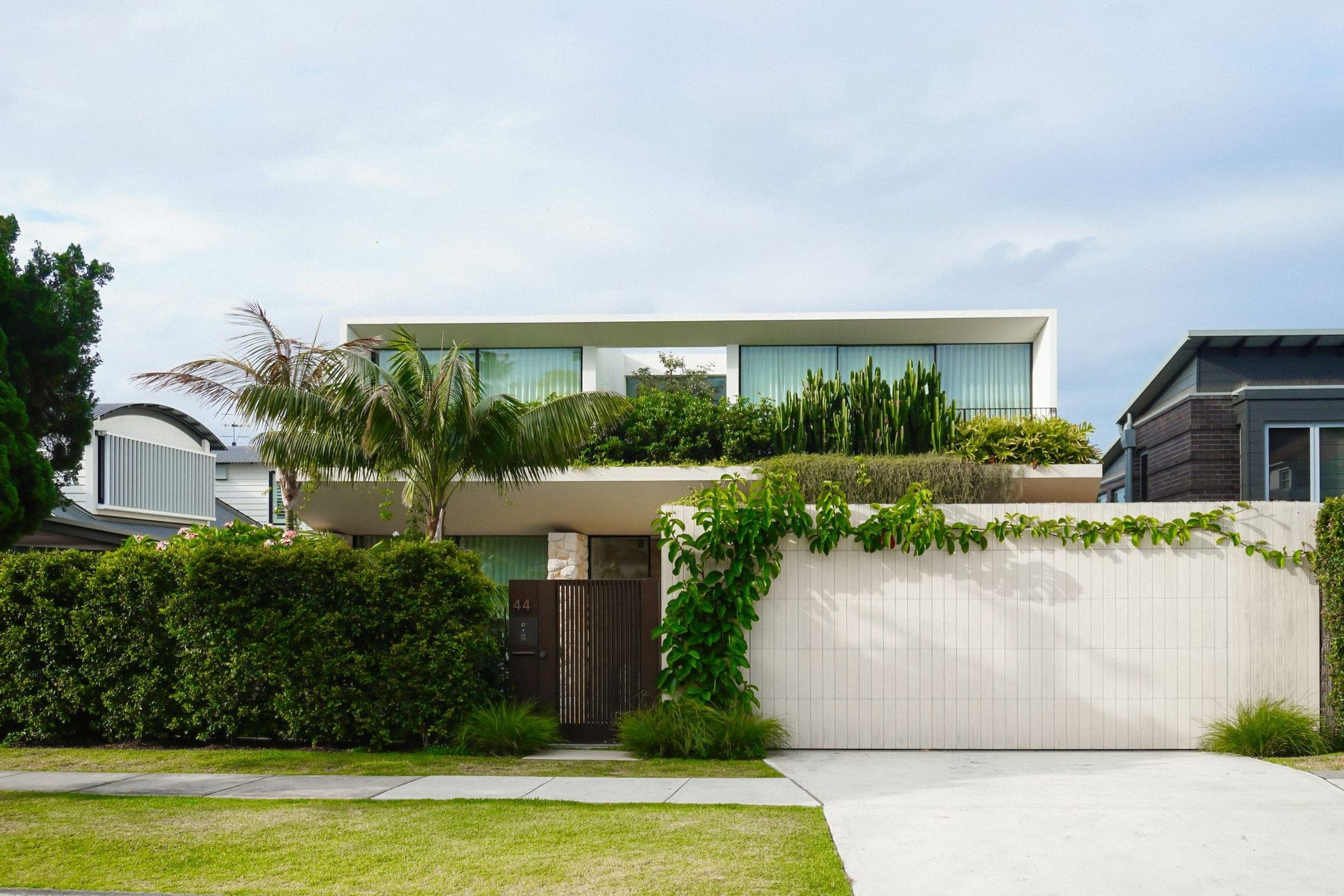 Two-storey rendered home with a timber-batten gate, palms and planted terraces from the street